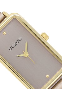 Montre rectangulaire en or avec un cadran gris brossé, des aiguilles dorées et un petit accent doré. Présente une couronne en or et un bracelet beige.