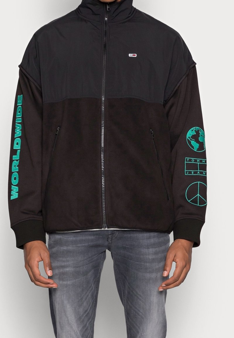 Černá bunda na zip s vysokým límcem. Má zelené detaily včetně nápisu "WORLDWIDE" a log na rukávech. Měkký materiál, volný střih.