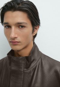 Giacca di pelle marrone con un colletto alto, texture liscia e una sottile trama. Primo piano del volto di un modello maschile, sfondo neutro.