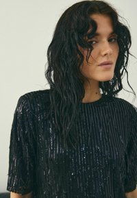 Schwarzes Pailletten-Oberteil mit strukturiertem Design, kurzen Ärmeln und lockerem Schnitt. Mit vertikalen und horizontalen Pailletten für einen schimmernden Effekt.