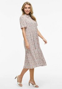 Kwiatowa sukienka midi z krótkimi rękawami, kołnierzykiem i marszczonym pasem. Jasne tło z czerwonym i niebieskim wzorem kwiatowym. Noszona z kremowymi sandałami na obcasie.