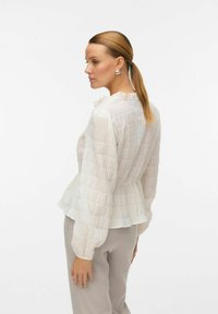 Vit texturerad blus med en volangkrage, långa puffärmar och en något peplumdesign, i kombination med ljusgrå byxor.