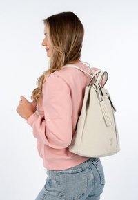 Mochila de cuero perforado en gris claro, con un acabado texturizado, que presenta correas en contraste y un zip lateral con un detalle de borlas.