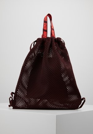 Donker kastanjebruine mesh rugzak met trekkoord, met rode en zwarte Nike-logo bandjes, tentoongesteld op een wit voetstuk tegen een lichtgrijze achtergrond.