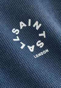 Valged "SAINT SALV" tähed, mis on paigutatud ringina tekstuurse tumesinise kangapõhja kohal, mille all on sõna "LONDON".