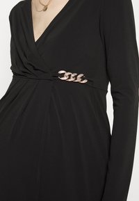 Vestido longo preto de mangas compridas com um decote em V profundo, apresentando um design torcido na cintura e um detalhe em corrente metálica com strass.