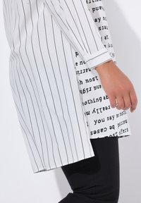 Camicia bianca a righe verticali nere, con pannello laterale contrastante di testo nero. Maniche lunghe con polsini abbottonati.