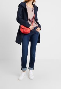 Marineblauer Kapuzenmantel über einem blumigen rosa Top, dunkle Jeans, die an den Säumen hochgekrempelt sind, weiße Sneakers und eine kleine rote Umhängetasche.