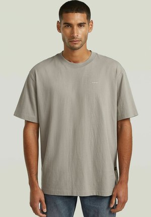 Homme aux cheveux courts et foncés portant un T-shirt ample gris clair et un jean bleu, debout devant un fond uni.