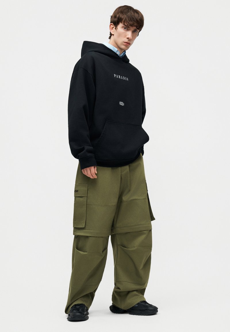 Joven con sudadera negra con capucha y texto "PARADIS" y pantalones cargo verde oliva de gran tamaño, posando frente a un fondo blanco liso.