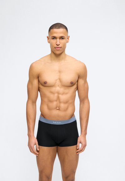 Μυώδης άνδρας με κοντά μαλλιά που στέκεται προς τα εμπρός φορώντας μαύρα μποξεράκια με γκρι ζώνη μέσης, μπροστά σε απλό ανοιχτό φόντο.