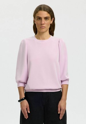 Kvinne med bølgete brunt hår iført en lys rosa topp med pufferm, svarte bukser og et svart armbånd, stående mot en ensfarget bakgrunn.