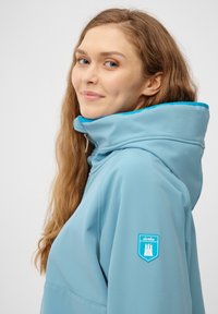 Ljusblå huva jacka med hög krage, kontrasterande teal-fodring och ett sköldpatch på vänster ärm med en tre-torn design.