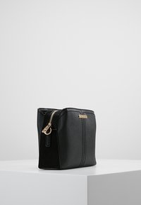 Svart kosmetikväska med en texturerad yta, guldfärgad dragkedja och en logotypplakett. Rektangulär form med minimalistisk design.