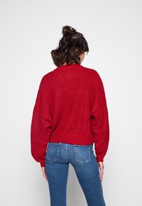 Donna con i capelli alla spalle, che indossa un maglione rosso spesso e jeans blu, in piedi con le spalle rivolte verso un fondo chiaro e uniforme.