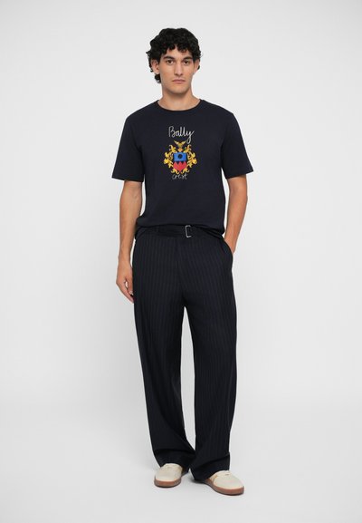 T-shirt in cotone blu navy con un design di stemma multicolore a contrasto, abbinato a pantaloni larghi a righe e scarpe di colore chiaro.