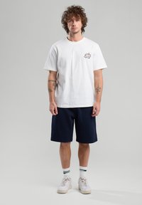 White cotton t-shirt with short sleeves and a small graphic, paired with navy shorts and white sneakers featuring green-striped socks.