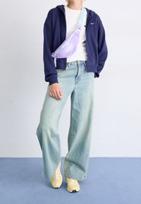 Lila und blaue Umhängetasche, getragen über einem weißen Shirt und einer navyblauen Kapuzenjacke, kombiniert mit hellblauen weit geschnittenen Jeans und gelben Sneakers.