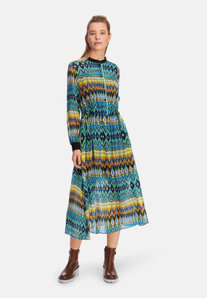 Langärmliges, gemustertes Kleid in lebhaftem Blau, Grün und Gelb mit taillierter Taille, versehen mit einem schwarzen Ausschnitt. Getragen mit braunen Knöchelstiefeln.