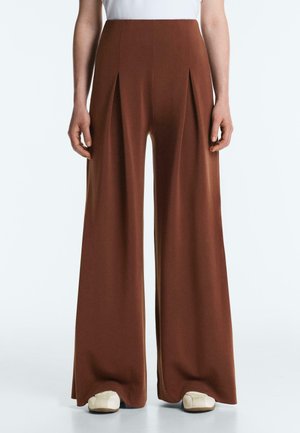 Pantaloni marroni a vita alta con gamba larga e pieghe frontali, indossati con scarpe bianche morbide e una maglia bianca infilata dentro.