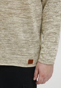 Pulover tricotat bej deschis, cu un model texturat, guler rotund și mâneci lungi. Prezintă o etichetă din piele maro la tiv.