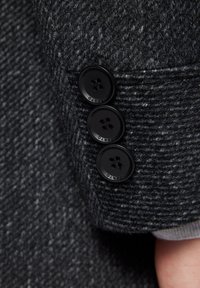 Szary materiał w wzór houndstooth na rękawie kurtki, z trzema czarnymi guzikami, każdy z czterema otworami, prezentujący teksturowany splot.