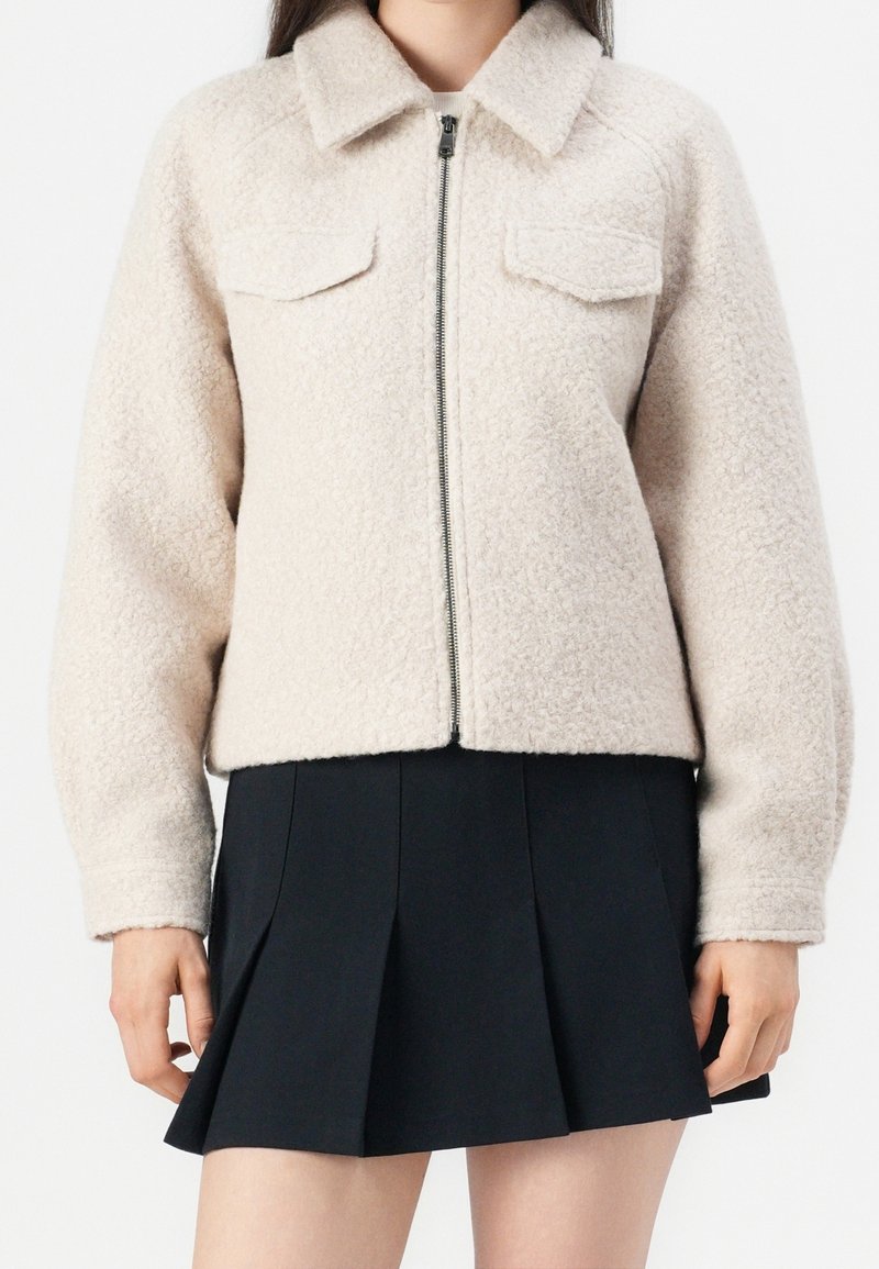 Femme portant une veste texturée crème avec une fermeture éclair à l’avant et des poches poitrine, associée à une mini-jupe plissée noire.