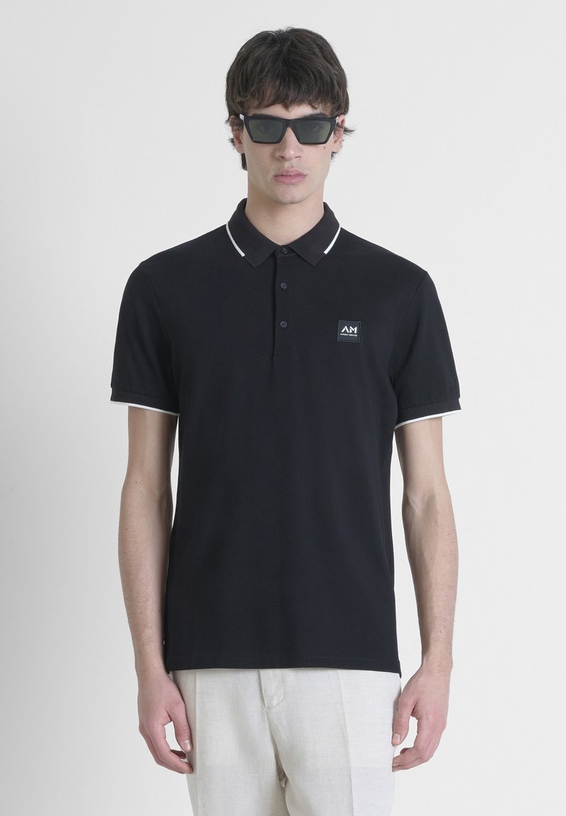 Zwarte polo van soepel materiaal, voorzien van een kraag met witte accenten en een klein logo-patch op de linkerborst. Model draagt een zonnebril.