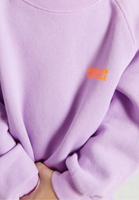 Fioletowy sweatshirt wykonany z miękkiego materiału, z ribbowanym dekoltem i mankietami, z pomarańczowym logo "AMERICAN VINTAGE" z przodu.