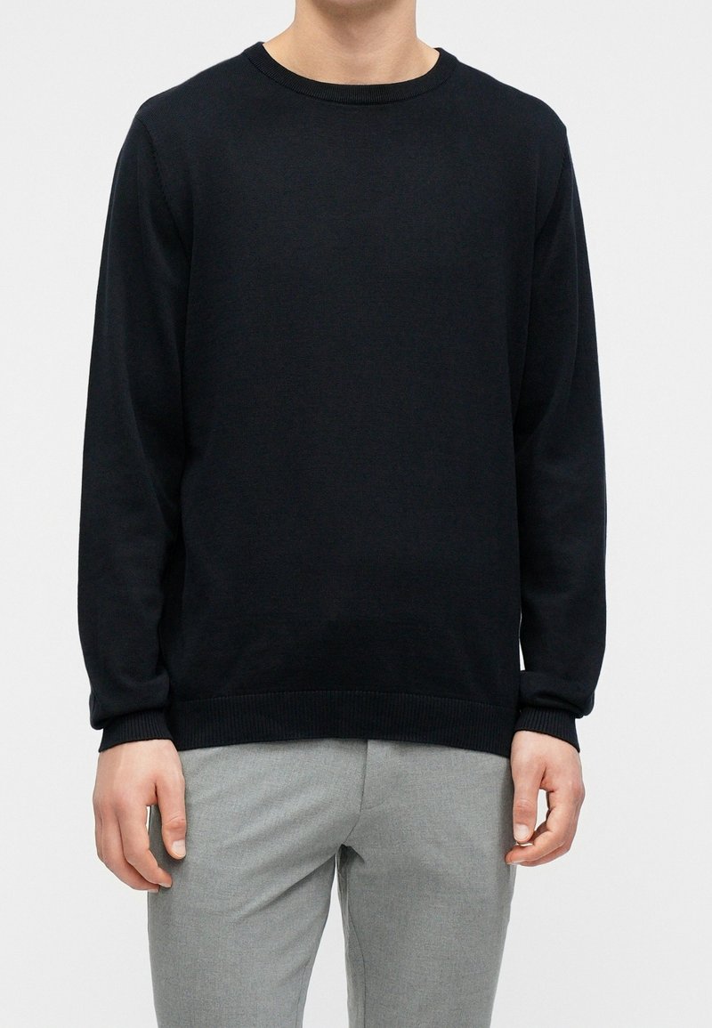 Homme portant un pull noir uni à manches longues et un pantalon gris clair, debout devant un fond neutre.