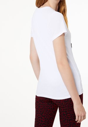 Donna con pelle chiara e capelli rossi, che indossa una semplice t-shirt bianca a maniche corte e pantaloni rossi con stampa leopardata, in piedi davanti a uno sfondo bianco.