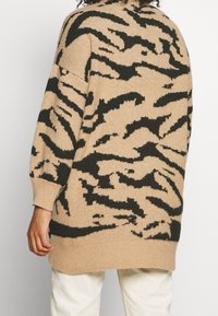 Suéter beige con un patrón de rayas de tigre negro. Corte holgado, hombros caídos y puños acanalados. Textura suave, termina por debajo de las caderas.