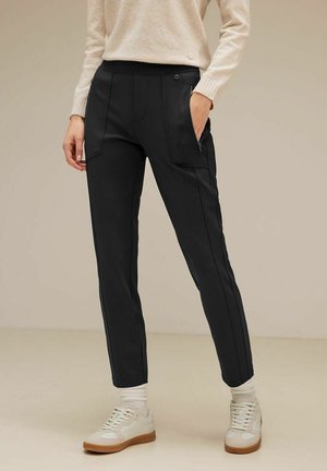 Schwarze schmal geschnittene Hosen aus weichem Material, mit zwei Reißverschlusstaschen und kontrastierenden Seitennähten. Kombiniert mit weißen Sneakers und einem cremefarbenen Pullover.