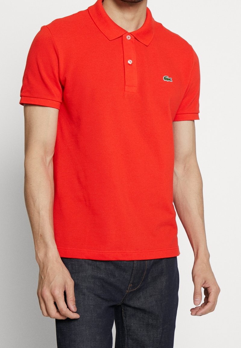 Homme portant un polo rouge vif à manches courtes avec un petit logo crocodile vert sur la poitrine et un jean bleu foncé, debout devant un fond blanc.
