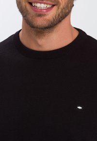 Czarny sweter z dzianiny z okrągłym dekoltem i ściągaczami na rękawach, z małym, białym logo na piersi. Gładka tekstura.