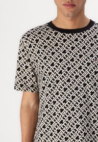 Μαύρη και άσπρη patterned μπλούζα με γεωμετρικό σχέδιο, κοντά μανίκια και μαύρη κλειστή λαιμόκοψη. Λεπτομέρεια του λογότυπου στο στήθος.