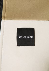 Columbia-märke på tyg, med en svart rektangulär etikett med vit logotyp. Den övre delen är tan, och den nedre delen är krämfärgad.