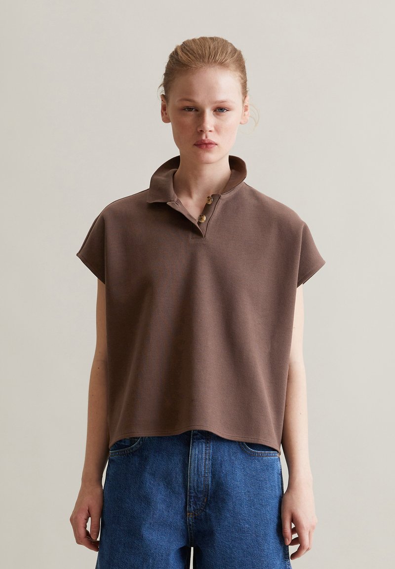 Braun das Poloshirt mit kurzen Ärmeln und Kragen, ausgestattet mit drei Knöpfen, aus strukturiertem Stoff. Kombiniert mit hoch taillierten blauen Jeansshorts.