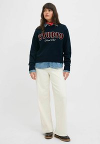 Jūras spēku svītra ar izšūtu "STUDIO Social Club", valkāta virs džinsa krekla. Saskaņota ar krēmkrāsas platām biksēm un brūnajām kurpēm.