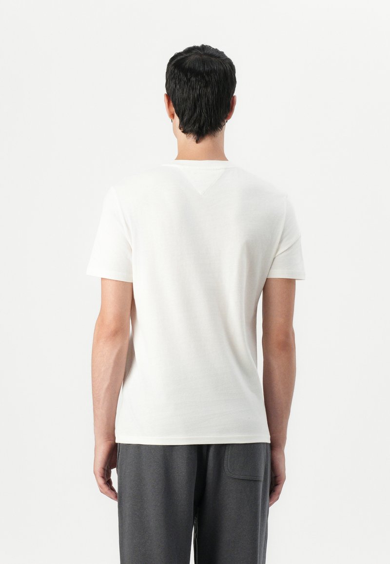 Camiseta blanca de algodón con mangas cortas, un cuello redondo clásico y un pequeño detalle en forma de V en la parte posterior. Llevada con pantalones de chándal grises.