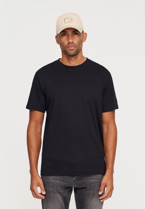 Black cotton t-shirt with a crew neck, worn with gray jeans. A beige cap with a logo is also visible. Simple design, casual fit.