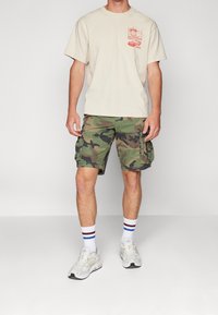 Mies, joka on pukeutunut kermaan sävyttyvään painokuvioiseen t-paitaan, vihreisiin maastokuvioisiin cargo-shortseihin, valkoisiin raidallisiin sukkiin ja valkoisiin tennareihin, seisomassa yksiväristä taustaa vasten.