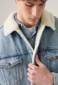 Denim jacket with a white sherpa collar, light blue color, chest pockets, metal buttons, and a red logo tag near the pocket.