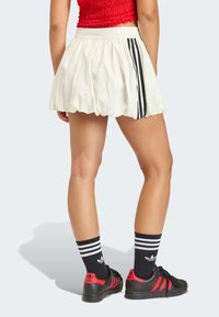 Bijela suknja trapezastog kroja s crnim prugama, uparena s crnim cipelama s crvenim detaljima i crnim čarapama do koljena s prugama. Glatka tekstura.