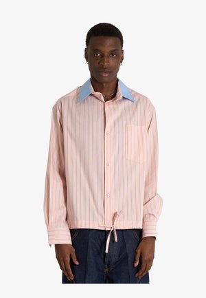 Uomo che indossa una camicia rosa a righe con bottoni, colletto blu e dettaglio cravatta all'orlo, abbinata a pantaloni blu scuro.