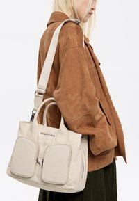 Sac fourre-tout beige avec double anses supérieures et une sangle amovible et réglable, comportant deux poches avant et un logo subtil. Texture lisse.
