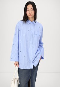 Chemise rayée bleu clair en tissu avec des accents carrés noirs. Elle a un col, des manches longues et une poche plaquée sur la poitrine.