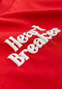 Punane puuvillane rõivaese, millel on reljeefsed valged tähed "Heart Breaker". Tekstuur on sile ja kaelus soonikkudele iseloomulik.