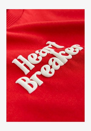 Prenda de algodón roja con texto en relieve blanco que dice "Heart Breaker". La textura es suave con un cuello acanalado.