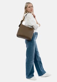 Donna che indossa una camicia bianca, jeans blu, sneakers bianche e che porta una grande borsa a tracolla marrone sulla spalla destra, su uno sfondo semplice.